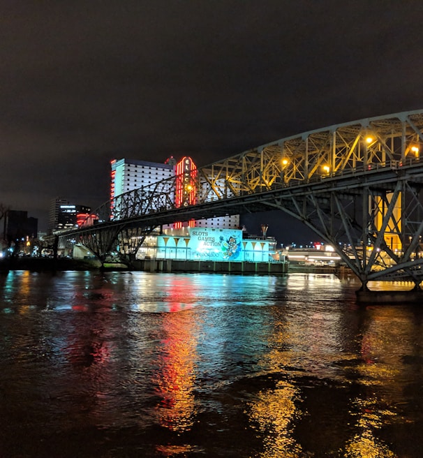 A brightly lit casino with neon signs and red lights is reflected in a river. A bridge stretches across the water, with an urban skyline visible in the background against an overcast night sky.