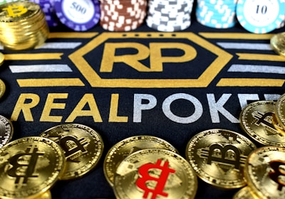 Stacks of poker chips in various colors are arranged in the background, while in the foreground, there are numerous golden coins featuring the Bitcoin symbol. The text 'REALPOKER' is prominently displayed in gold and silver lettering on a dark background.