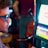 A man wearing sunglasses is intently focused on a slot machine, with colorful lights and graphics illuminating the screen. The scene is set indoors, likely in a casino or gaming area. His attire includes a patterned shirt and a blazer.