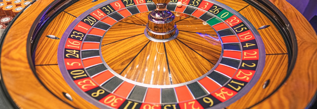 A roulette wheel with alternating red and black numbered pockets on a wooden platform. The wheel contains a small white ball resting in one of the pockets, indicating gameplay. Surrounding the wheel are stacks of colorful poker chips, suggesting a casino environment.