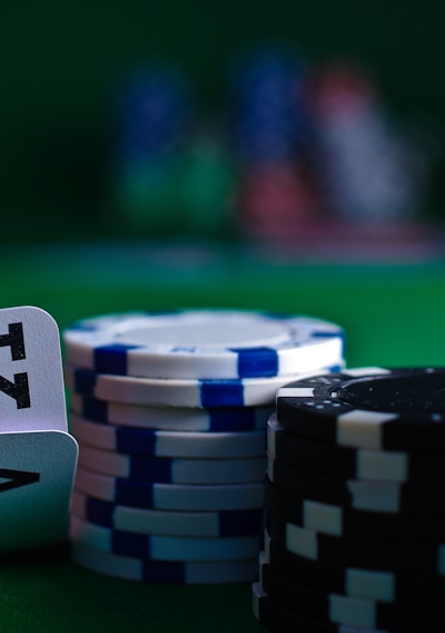 A casino scene featuring two playing cards, the ace and king of clubs, partially concealed by a player's hand. Nearby, there are stacks of multicolored poker chips on a green felt table, with blurred colorful background elements suggesting other chip stacks or cards.